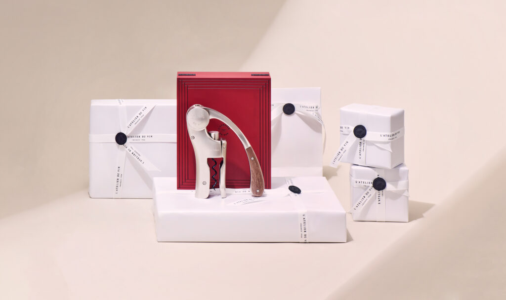 Luxuriöser Œno Motion Trésor Korkenzieher präsentiert vor seinem roten Etui, umgeben von elegant verpackten Geschenkboxen aus weißem Papier mit Bändern und personalisierten Siegeln.