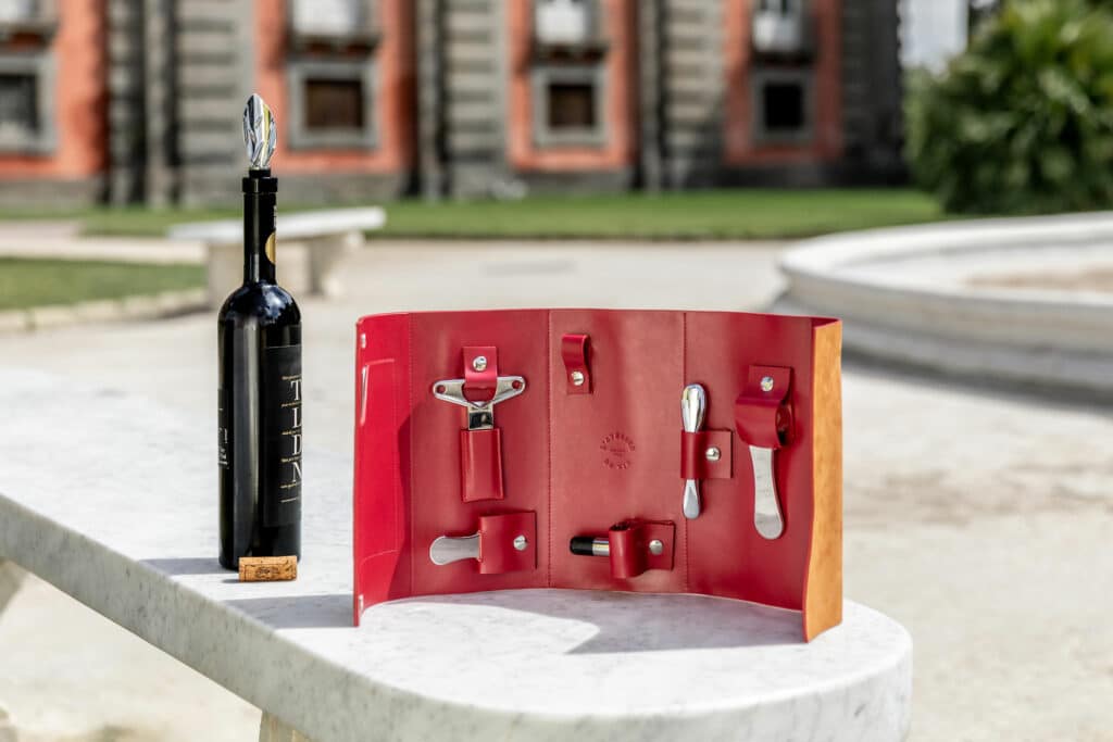 Rote Nomadentasche aus Leder, geöffnet auf einer weißen Marmorfläche, enthält Weinaccessoires — Korkenzieher, Folienabschneider und Servicetools — neben einer Weinflasche, im Freien mit Gebäuden und Grünflächen im Hintergrund.