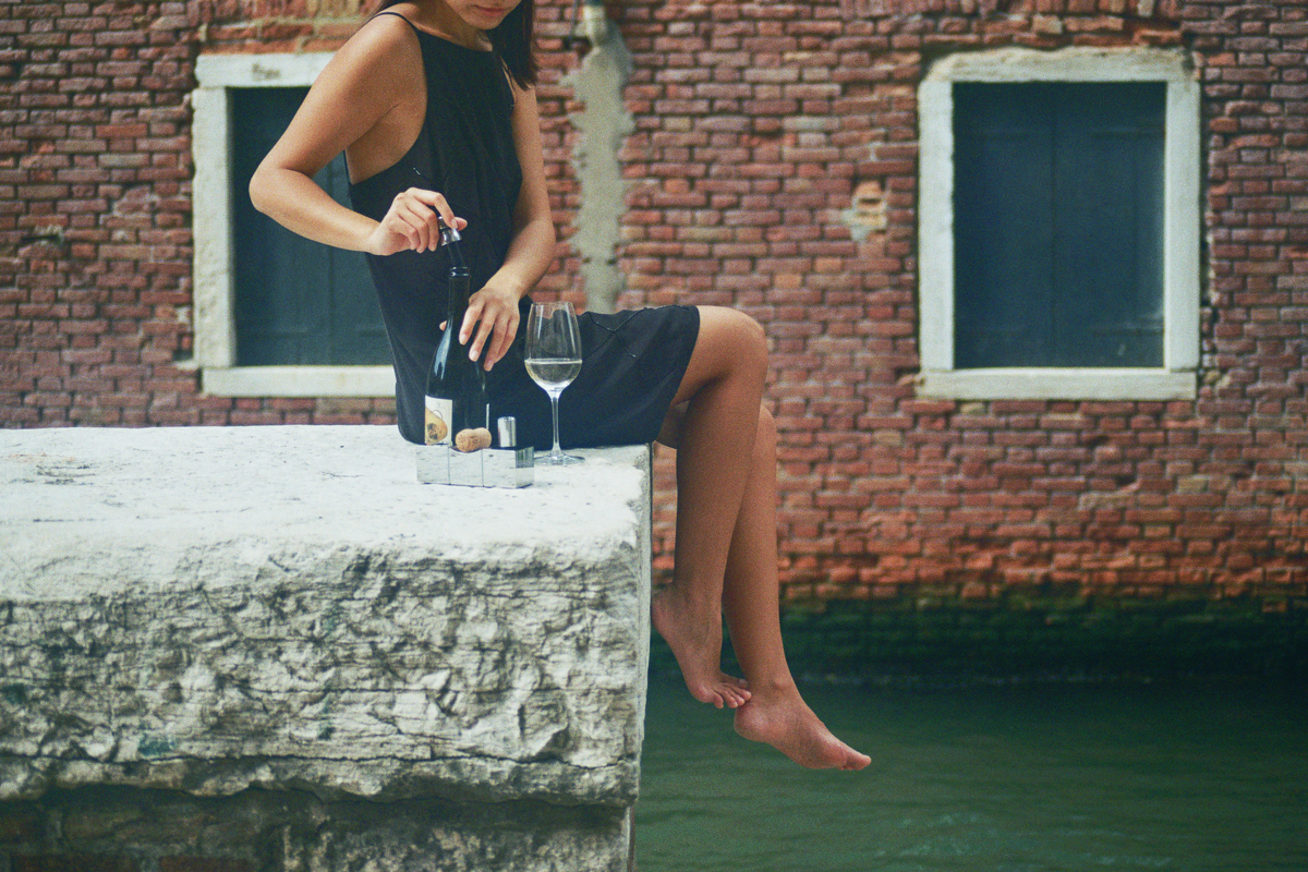 Woman in black dress sitting on a Venice quay, placing a Bubble Cork stopper from L'Atelier du Vin on a sparkling wine bottle