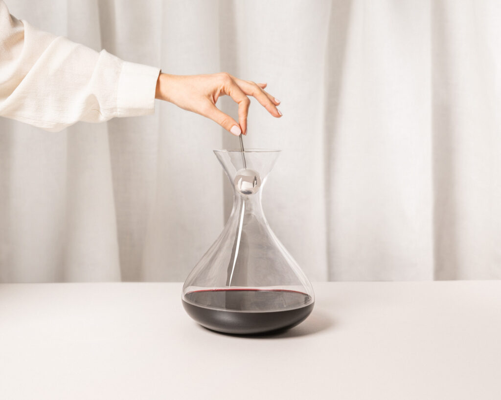 Main tenant un developer boule au-dessus d'une carafe en verre contenant du vin rouge, posée sur une surface claire avec un rideau blanc en arrière-plan