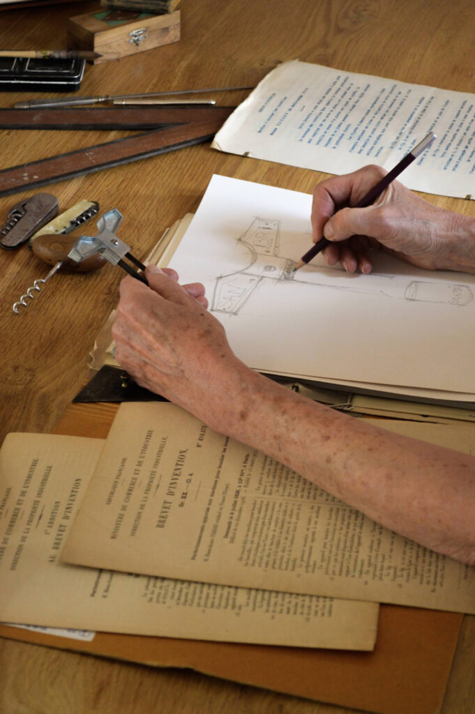 Personne réalisant un croquis technique du bilame à la main sur une feuille de papier, entourée d'outils de mesure, d'un document ancien de brevet d'invention et de tire-bouchons sur une table de travail.