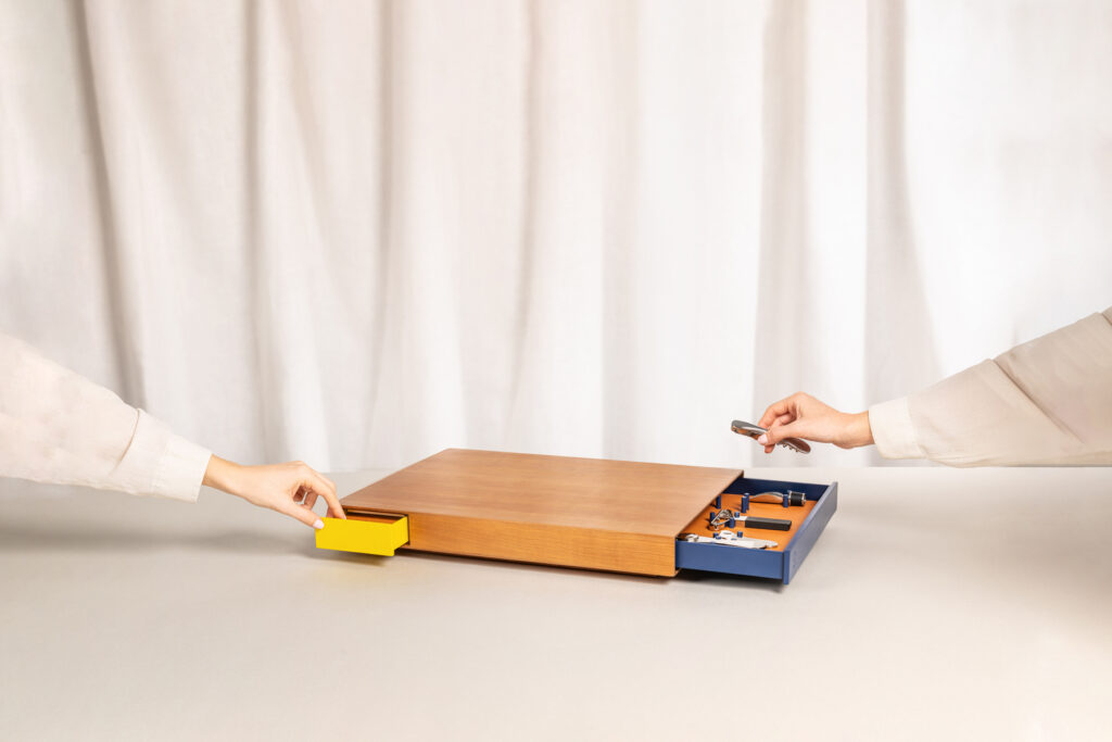Plateau en bois design avec deux tiroirs coulissants colorés — un jaune et un bleu — contenant des accessoires pour le vin comme un tire-bouchon, manipulé par deux mains, illustrant l’usage pratique et élégant de l’objet.