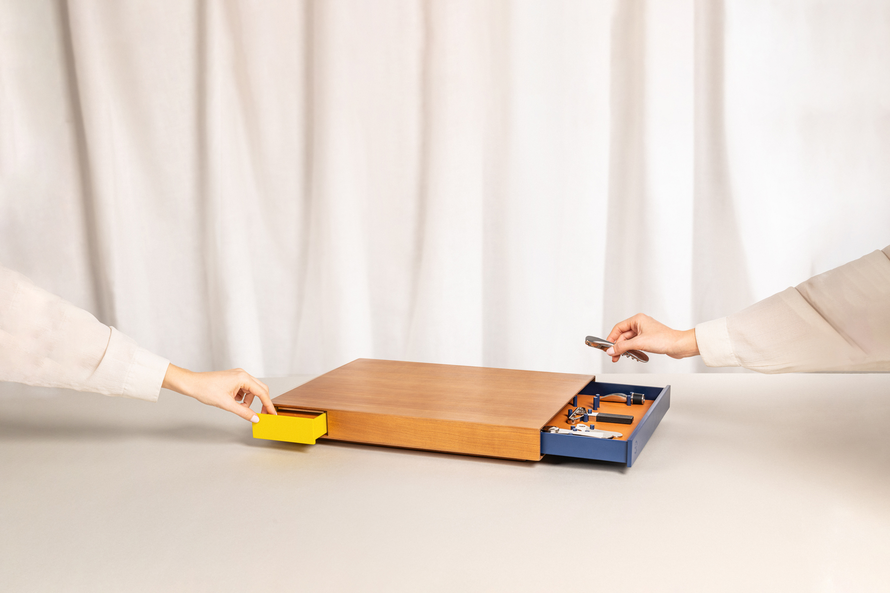 Plateau en bois design avec deux tiroirs coulissants colorés — un jaune et un bleu — contenant des accessoires pour le vin comme un tire-bouchon, manipulé par deux mains, illustrant l’usage pratique et élégant de l’objet.