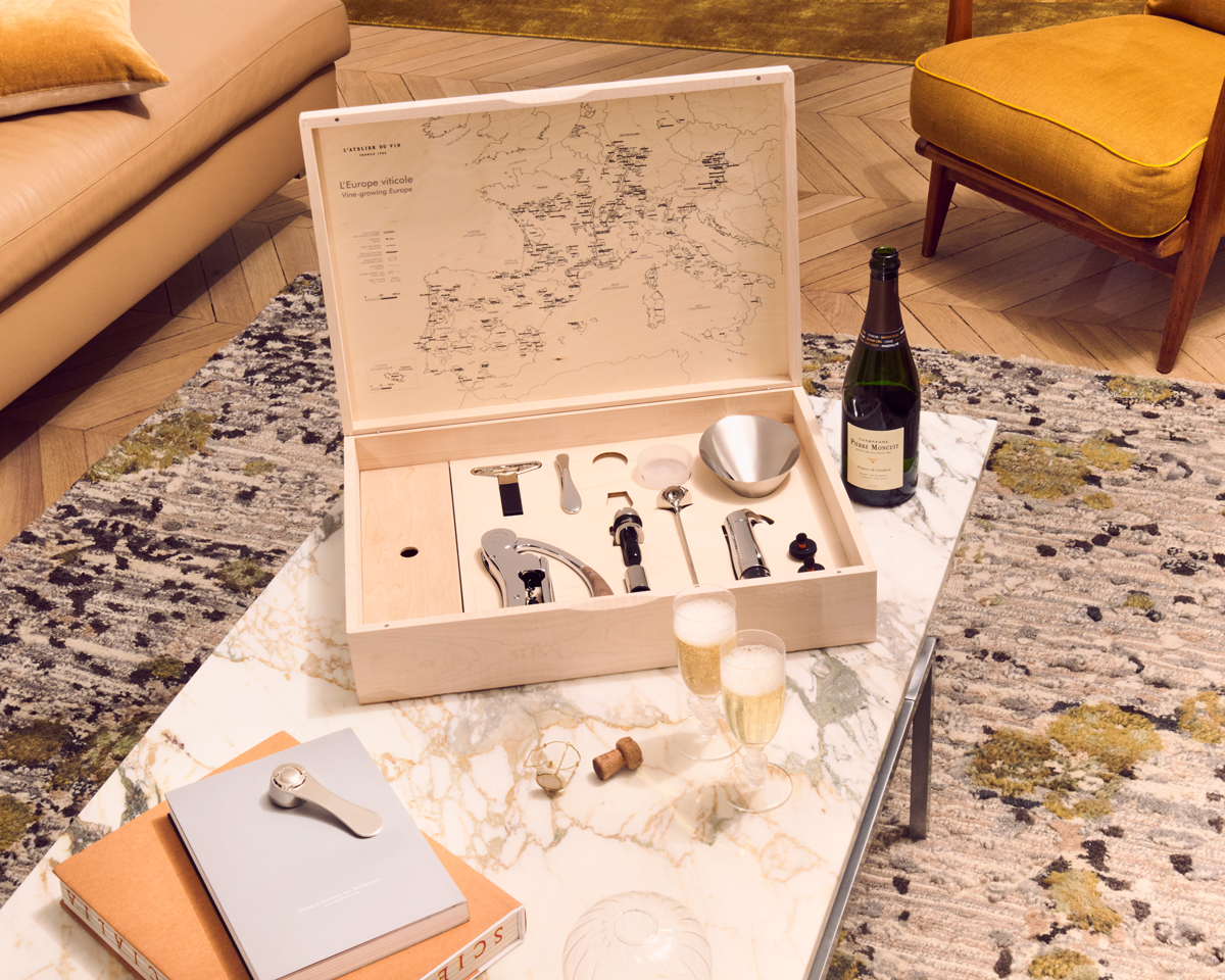 Coffret oenologique Oeno Box Connoisseur ouvert, présenté sur une table en marbre, dévoilant ses accessoires œnologiques haut de gamme, avec une bouteille de vin et deux verres de champagne dans un intérieur chaleureux et raffiné.