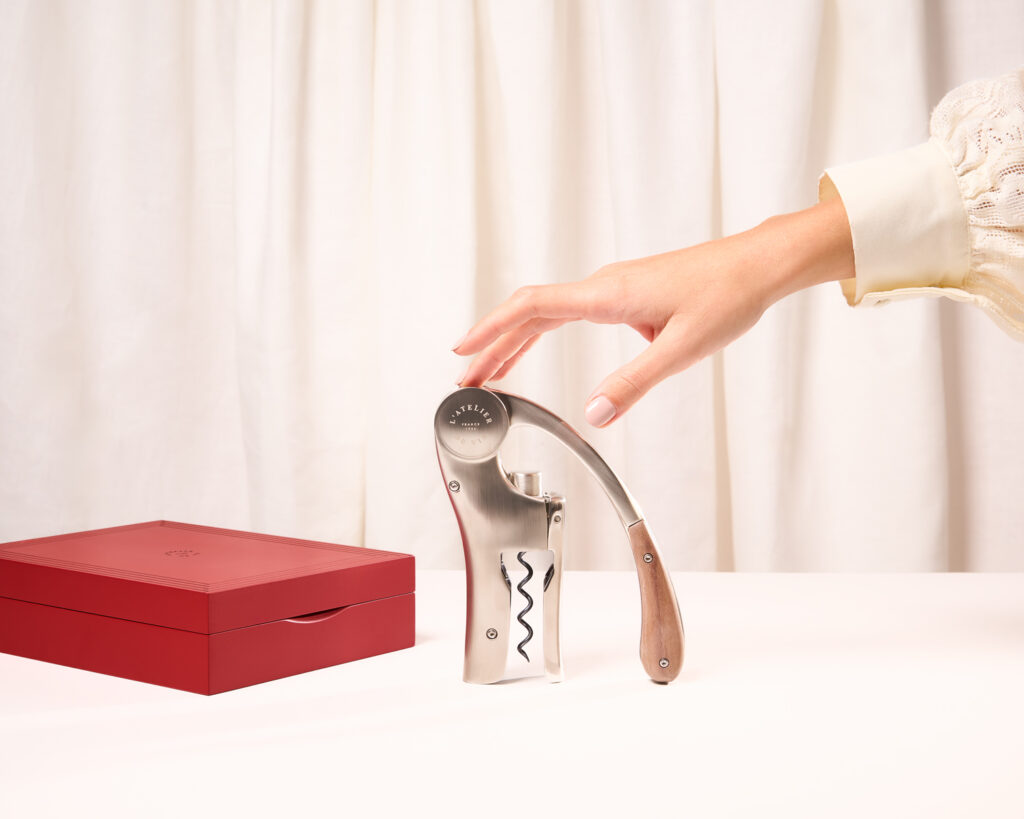 Main s’approchant d’un tire-bouchon avec manche en bois, posé verticalement sur une surface blanche, à côté d’un coffret rouge, avec rideaux blancs en arrière-plan.