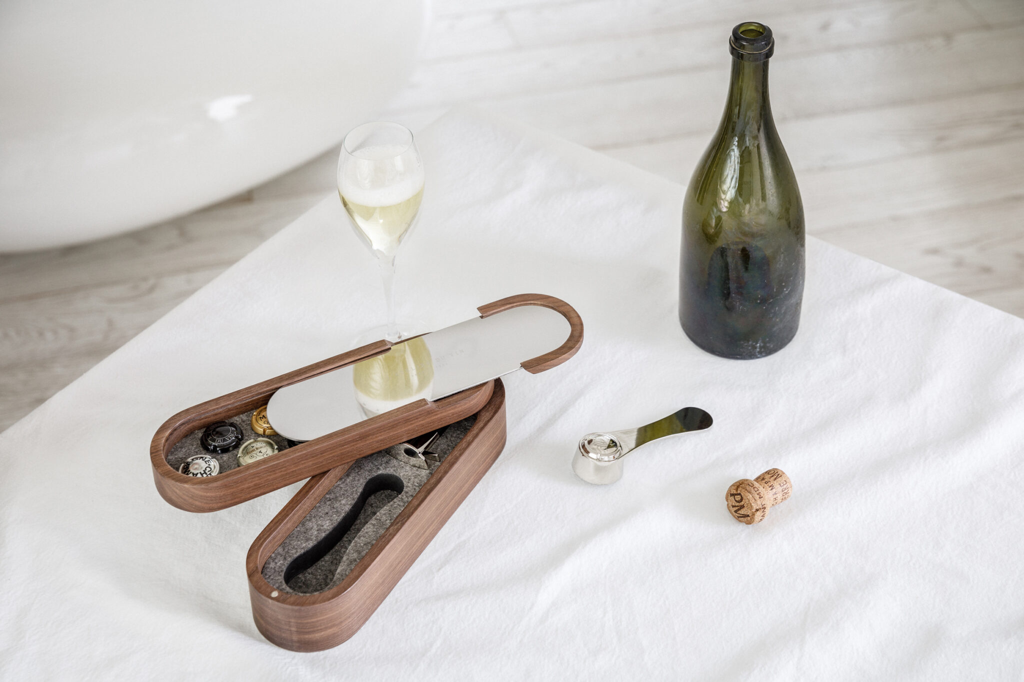 Holzkoffer mit Spiegeldeckel, geöffnet auf einer weißen Tischdecke, enthält Zubehör für den Service von Schaumwein — Museletzange, Konservierungsverschluss, Platz für Kapselsammlungen — begleitet von einer geöffneten Flasche Schaumwein, einem gefüllten Glas und einem Verschluss.