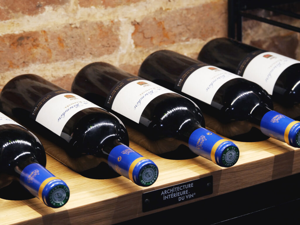 Bouteilles de vin disposées horizontalement sur une tablette en bois ajourée signée Architecture Intérieure du Vin, devant un mur en briques.