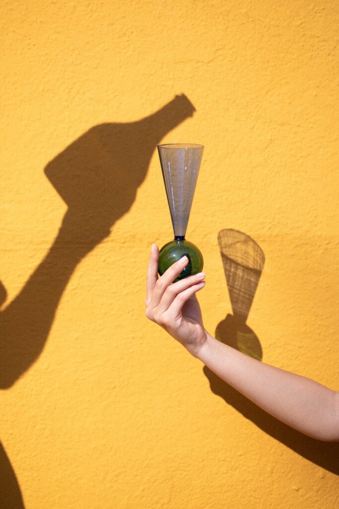 Main tenant une flûte Bubbles Celebration avec base sphérique verte, projetant une ombre sur un mur jaune
